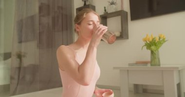 Pembe yoga kıyafetli kadın yoga çalışmasını iyi aydınlatılmış, minimalist bir oturma odasında tamamladıktan sonra bir yudum su alıyor. Sahnede rahatlama, susuzluk ve dinginlik vurgulanıyor.