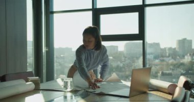 Odaklanmış Asyalı bir kadın güneşli bir ofiste mimari planlar üzerinde çalışıyor, bir dizüstü bilgisayarla ve masanın üzerinde yuvarlanmış planlarla, büyük pencerelerden panoramik bir şehir manzarasına bakıyor..