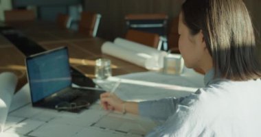 Günışığı altındaki modern bir çalışma alanında odaklanmış bir mimar, geniş bir konferans masasının taslaklarını analiz ediyor, taslak araçları, dizüstü bilgisayarı ve yuvarlanmış planlarıyla, yaratıcılık ve hassasiyet sergiliyor..