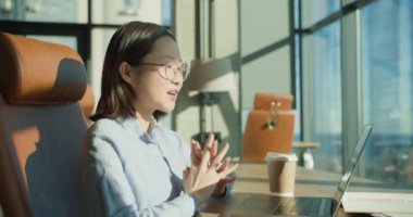 Gözlüklü mutlu bir genç kadın, parlak ve modern bir ofiste oturuyor, bir video görüşmesi sırasında el sallıyor. Çalışma sahasında bir dizüstü bilgisayar, kahve fincanı ve doğal ışık, pozitiflik ve profesyonellik yayılıyor