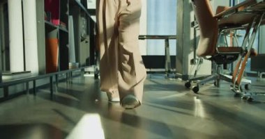 Aydınlık ve modern bir ofis alanında yürüyen bir kadının yakın çekimi. Sahne bej pantolonları, beyaz spor ayakkabıları ve doğal ışıklandırması olan şık bir çalışma alanını ön plana çıkarıyor.. 
