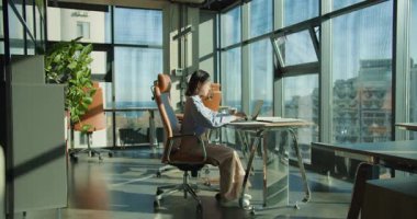 Profesyonel bir kadın, güncel, güneşli bir ofiste elinde bir kahve fincanı tutarak bilgisayarla masasında oturuyor. Geniş bir çevrede şehir manzaralı büyük pencereler vardır..