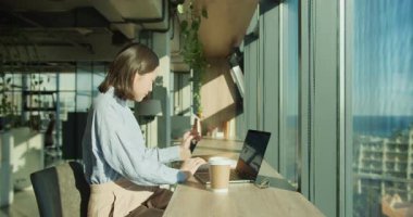 Mavi gömlekli bir iş kadını parlak ve modern bir ofiste otururken bir defter okumaya odaklanır. Çalışma alanında büyük pencereler, yeşillik, bir kahve fincanı ve bir dizüstü bilgisayar bulunuyor..