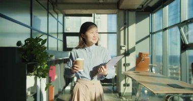 Genç bir kadın, elinde kahve fincanı ve not panosuyla modern bir ofiste kendinden emin bir şekilde duruyor. Büyük pencereler, kapalı alandaki bitkiler ve şık mobilyalarla tamamlanan güneş ışığının boşluğu doldurmasına olanak sağlıyor.