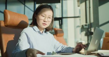 Gözlüklü ve açık mavi gömlekli odaklanmış genç profesyonel bir kadın modern bir ofiste bir masada oturuyor. Dizüstü bilgisayarda çalışıyor ve gün ışığında belgeleri inceliyor. Masada kahve ve çizelgeler var.