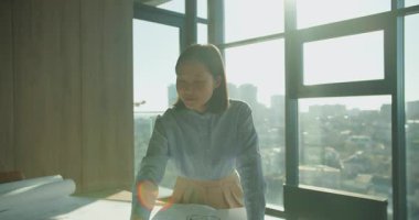 Profesyonel bir kadın elinde dizüstü bilgisayarla masanın üzerine eğiliyor. Etrafı belgelerle çevrili ve arka planda parlak bir şehir manzarası var. Sahne odak, yaratıcılık ve modern çalışma kültürünü yansıtıyor..