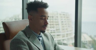 Çağdaş bir ofiste oturan, takım elbiseli, odaklanmış bir işadamı, büyük cam pencereli. Görüntü profesyonelliği, güveni ve kurumsal ortamı vurguluyor.