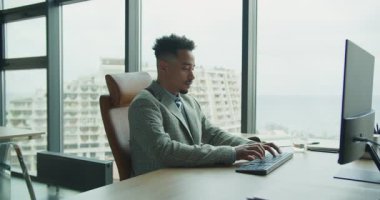 Terzi kıyafeti giymiş profesyonel bir işadamı şehrin panoramik manzaralı parlak, modern bir ofisinde klavyeye yazı yazıyor. Görüntü profesyonelliği, odaklanmayı ve işyeri verimliliğini aktarıyor.