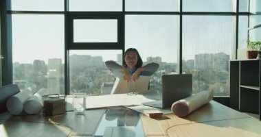 Yerden tavana pencereleri olan modern bir ofisin planlarını havaya fırlatan bir mimar. Masada proje araçları, bir dizüstü bilgisayar ve arkaplanda şehir manzaralı belgeler bulunur..