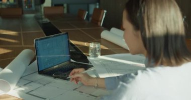 Profesyonel bir mimar, güneşli modern çalışma alanındaki bir dizüstü bilgisayarın dijital planlarını inceliyor. Şık bir konferans masasının üzerinde yuvarlanmış planlar ve çizim araçları var.