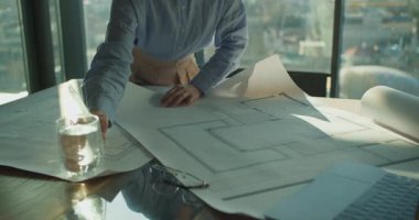 Bir masanın üzerine yayılmış mimari planların yakın plan görüntüsü, profesyonel bir el, doğal ışık ve şehir manzarası içeren parlak bir ofis ortamında tasarımı denetliyor..