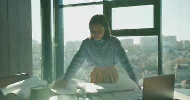 Şehir manzaralı, tasarım araçları, yuvarlanmış planlar ve ahşap masadaki bir dizüstü bilgisayarla çevrili modern bir ofisin inşaat planlarını dikkatle analiz eden bir mimar.