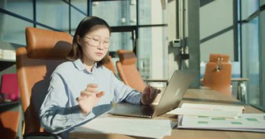 Çağdaş bir ofis ortamında masasında önemli belgeleri analiz eden gözlüklü genç bir iş kadını. Sıcak güneş ışığı, büyük cam pencerelerden akarak onun konsantrasyonunu vurguluyor.