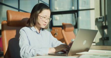 Gözlüklü profesyonel bir genç kadın, modern bir ofiste dizüstü bilgisayarıyla çalışıyor. Çalışma alanında deri bir sandalye, kahve fincanı ve verimli bir çalışma için düzenli notlar yer alıyor.