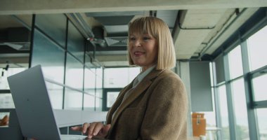 Modern bir ofiste dizüstü bilgisayarıyla çalışan kendinden emin bir iş kadını. Görüntü, onun kurumsal bir ortamda odaklanma, üretkenlik ve profesyonel davranışını vurguluyor..