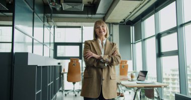 Modern bir ofiste kendine güvenen olgun bir iş kadını. Kollarını kavuşturup profesyonelliği ve liderliği yayıyor. Parlak çalışma alanı kurumsal ortamı yansıtıyor..