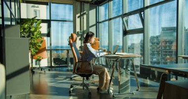 Profesyonel bir kadın, güncel, güneşli bir ofiste elinde bir kahve fincanı tutarak bilgisayarla masasında oturuyor. Geniş bir çevrede şehir manzaralı büyük pencereler vardır..