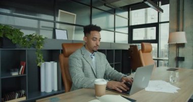 Gösterişli ofis ortamında profesyonel bir işadamı dizüstü bilgisayarda daktilo kullanıyor. Çalışma alanı, üretken ve modern bir atmosfer yaratan doğal ışık, grafikler ve bir kahve fincanı içeriyor..