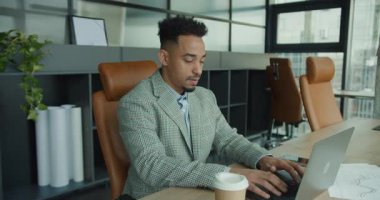 Özel dikim takım elbiseli profesyonel bir işadamı çağdaş bir ofiste dizüstü bilgisayarda yazı yazıyor. Masadaki bir kahve fincanı ve grafikler üretkenlik sahnesine ve modern bir iş yerine odaklanmaya katkı sağlar..