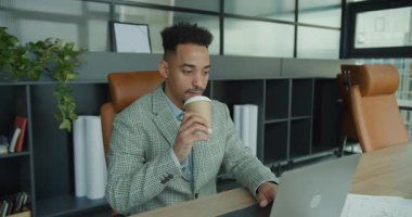 Modern bir ofiste dizüstü bilgisayar üzerinde çalışırken şık takım elbiseli profesyonel bir işadamı bir fincan kahve molası verir. Ayar vurguları odak, rahatlama ve işyeri verimliliği.