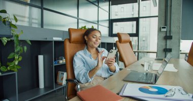 Mavi gömlekli odaklanmış genç bir kadın masasında oturuyor, parlak, modern bir ofiste, telefonla konuşuyor ve belgelerini gözden geçiriyor, bitkiler, çizelgeler, dizüstü bilgisayarlar ve parlak deri koltuklar ile çevrili.