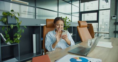 Mavi tişörtlü mutlu bir kadın video görüşmesi sırasında akıllı telefonuna el sallıyor. Elinde laptop, belgeler ve bitkilerle dolu modern bir ofiste bir bardak suyla bir masada oturuyor.
