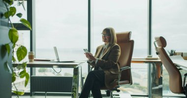 Modern bir ofiste akıllı telefon kullanan rahat bir iş kadını büyük cam pencereli ve arka planda şehir ve deniz manzaralı bir ofis. Yüksek kalite fotoğraf