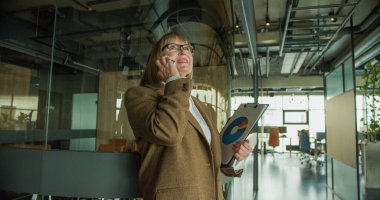 Modern bir ofiste odaklanmış bir iş kadını, elinde veri tablosu olan bir pano tutarken bir telefon görüşmesi yapıyor, güven ve profesyonellik gösteriyor. Yüksek kalite fotoğraf