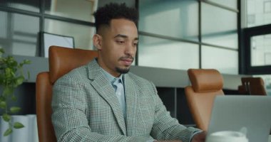 Kareli takım elbise giyen kendine güvenen bir işadamı, şık ve modern bir ofis alanında çalışırken masasında sıcak bir gülümsemeyle oturuyor. Çevre yeşillik, raflar ve yumuşak doğayı içerir.