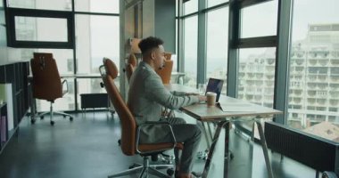 Özel dikim takım elbiseli profesyonel bir işadamı, çağdaş açık bir ofiste dizüstü bilgisayar üzerinde çalışıyor. Ayarlarda doğal ışık, büyük pencereler ve masanın üzerinde bir kahve fincanı yer alıyor.