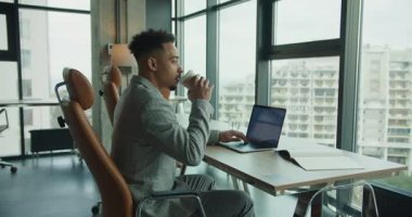 Şık takım elbiseli profesyonel bir işadamı bir dizüstü bilgisayar üzerinde çalışıyor ve parlak, açık bir ofiste kahvesini yudumluyor. Çalışma alanı doğal ışık, büyük pencereler ve odak noktasını vurgulayan bir şehir manzarası içerir.