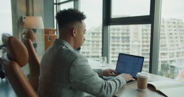 Kareli takım elbiseli profesyonel bir işadamı, parlak ve modern bir ofiste dizüstü bilgisayarına odaklanır. Çalışma alanı bir kahve fincanı, açık bir defter ve çarpıcı bir şehir manzarası içeriyor.