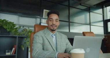 Şık bir damalı takım giyen profesyonel bir işadamı, modern bir ofiste dizüstü bilgisayarı üzerinde çalışırken derinden odaklanır. Modern çalışma alanı temiz hatlar, yeşillik ve doğallık içerir.