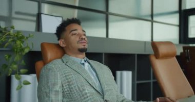 Şık damalı takım elbiseli kendine güvenen bir işadamı, şık, modern çalışma alanında ergonomik ofis sandalyesine yaslanır. Arkaplanda çağdaş dekor ve yeşillik yer alıyor, yansıtıcı