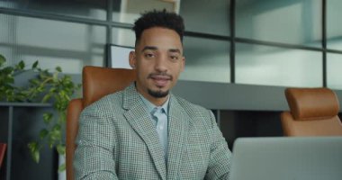 Kareli takım elbise giyen neşeli bir işadamı çağdaş bir ofis alanında güvenle duruyor. Büyük pencereler, profesyonel ve modern bir atmosfer oluşturan bir şehir manzarası sergiliyor. Yüksek