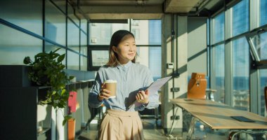 Genç bir kadın, elinde kahve fincanı ve not panosuyla modern bir ofiste kendinden emin bir şekilde duruyor. Büyük pencereler, kapalı alandaki bitkiler ve şık mobilyalarla tamamlanan güneş ışığının boşluğu doldurmasına olanak sağlıyor.