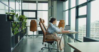 Modern profesyonel bir kadın, şık, parlak bir ofiste dizüstü bilgisayarla çalışıyor. Şehir manzaralı geniş pencereler, bitkiler ve çağdaş mobilyalar var. Yüksek kalite fotoğraf