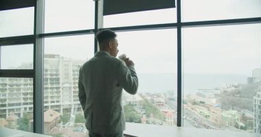 Kareli takım elbiseli bir işadamı büyük bir ofis penceresinin yanında elinde bir fincan kahve tutarken şehir manzarasına ve okyanus manzarasına bakıyor. Modern, endüstriyel ofis kurulumu odak noktalarını vurguluyor