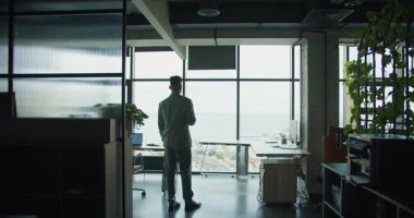 Kareli takım elbiseli bir profesyonel geniş ofis pencerelerinin yanında duruyor, şehre ve okyanusa bakıyor. Endüstriyel ofis tasarımı doğal ışık ile modern, yansıtıcı bir atmosfer yaratır. Yüksek