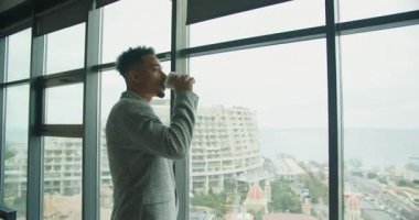 Kareli takım elbiseli bir işadamı, şehir manzarası ve deniz manzaralı büyük bir pencerenin yanında dururken kahve içer. Modern ofis sakin bir ortam yaratır..
