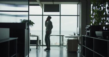 Modern bir ofis alanında yürüyen ve konuşan bir iş adamı. Siluet, arka planda şehir ve deniz manzaralı büyük pencereler tarafından çerçevelenmiştir. Yüksek kalite 4k görüntü