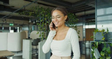 Parlak, modern bir ofiste, yeşil, gösterişli mobilyalar ve doğal ışık ile çevrili bir telefonda gülümseyen profesyonel kadın güven ve neşe saçıyor. Yüksek kalite fotoğraf