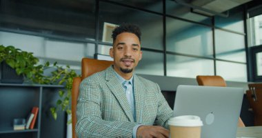 Kareli takım elbise giyen kendine güvenen bir işadamı, şık ve modern bir ofis alanında çalışırken masasında sıcak bir gülümsemeyle oturuyor. Çevre yeşillik, raflar ve yumuşak doğayı içerir.