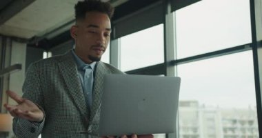 Modern bir ofisteki bir işadamı sanal bir tartışmaya girişir ve dizüstü bilgisayarına doğru hareket eder. Arka plandaki büyük pencereler profesyonel ve açık bir ortam sağlıyor. Yüksek kalite 4k görüntü