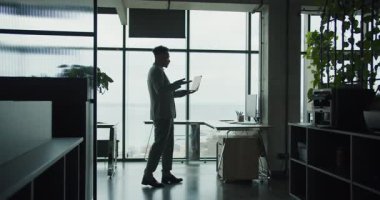 Çağdaş bir ofiste dizüstü bilgisayar kullanan bir iş adamının silüeti. Yerden tavana pencereler, şehir manzarası ve deniz manzarası. Mekan modern ve profesyonel. Yüksek kalite 4k görüntü