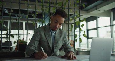 Profesyonel bir işadamı, geniş pencereleri, yeşilliği ve doğal ışığı olan modern bir ofisteki belgelere odaklanır. İş, planlama ve üretkenlik temaları için ideal. Yüksek kalite 4k