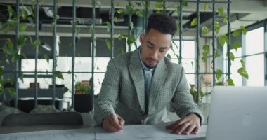 Bir mimar, çevresi yeşilliklerle çevrili çağdaş bir çalışma alanında ayrıntılı planlar çizmeye odaklanır. Bu görüntü yaratıcılığı, hassasiyeti ve profesyonel atmosferi yansıtıyor. Yüksek kalite 4k