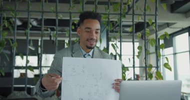 Modern bir çalışma alanındaki neşeli bir profesyonel sanal toplantı sırasında detaylı bir kat planını açıklar. Arka planda canlı bir yeşillik ve yeni bir ambiyans var. Yüksek kalite 4k görüntü