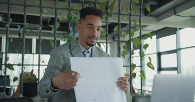 Kareli takım elbiseli kendine güvenen bir işadamı sanal sunum sırasında detaylı bir teknik çizim yapıyor. Şık ofis arka planı yeşillikle süslenmiş. Yüksek kalite 4k görüntü