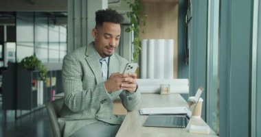 Ekose takım elbiseli bir işadamı modern bir ofis masasında otururken akıllı telefonunu kullanır. Çalışma alanı bir dizüstü bilgisayar, kahve fincanı ve dokümanlar içeriyor.